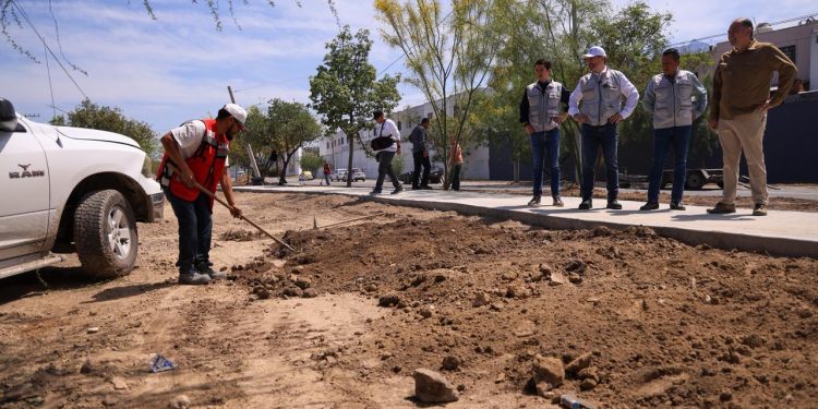 Construction Worker in a Red Safety Vest Shovels Dirt Beside a White Pickup While Five Colleagues Watch from the Sidewalk. - Hola Mty