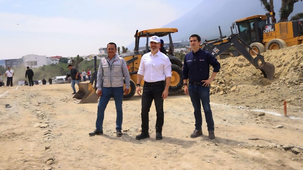 Three Men Stand on a Dusty Construction Site with Heavy Machinery Behind Them, Posing for a Photo During a Project Visit. - Hola Mty