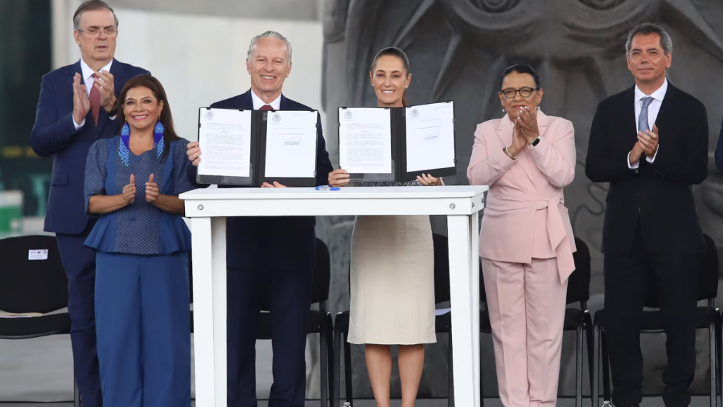 Six Officials in Formal Attire Stand Behind a White Table As Two Center Figures Display Signed Documents, Others Applaud, Indicating a Signing Ceremony. - Hola Mty