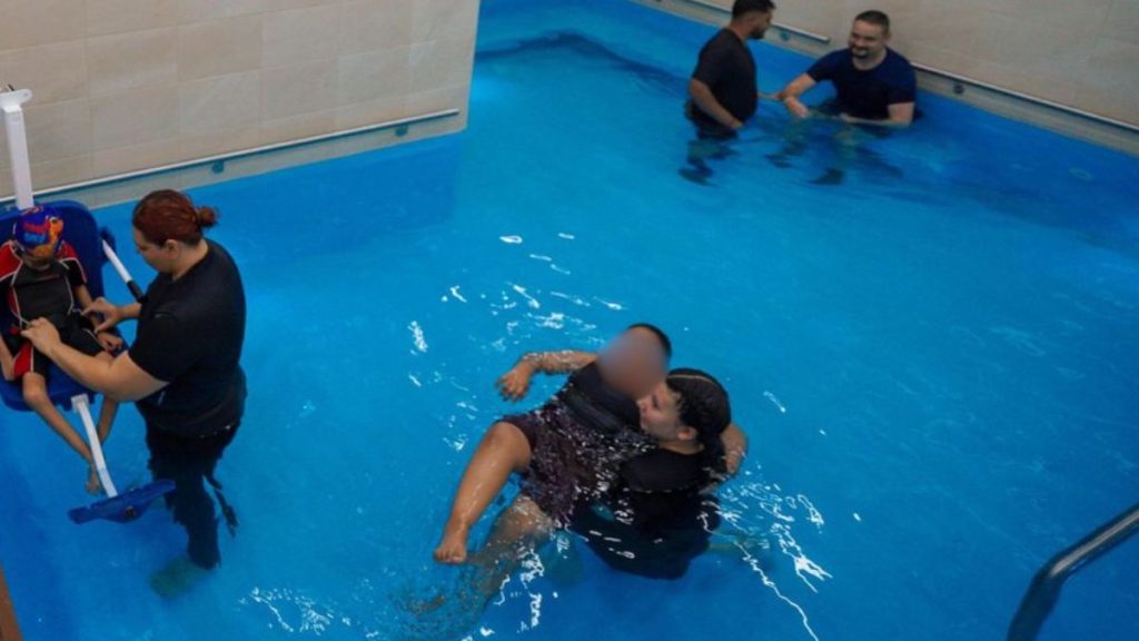 Instructor Assists a Child in a Flotation Chair at Pool Edge with Another Adult Nearby in the Water in a Rehabilitation or Swimming Lesson Setting - Hola Mty