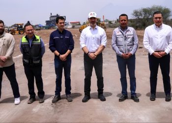 Six Men in Work Clothes Stand in a Row on a Dirt Construction Site, with Heavy Machinery in the Background. - Hola Mty