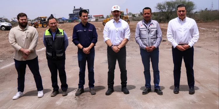 Six Men in Work Clothes Stand in a Row on a Dirt Construction Site, with Heavy Machinery in the Background. - Hola Mty
