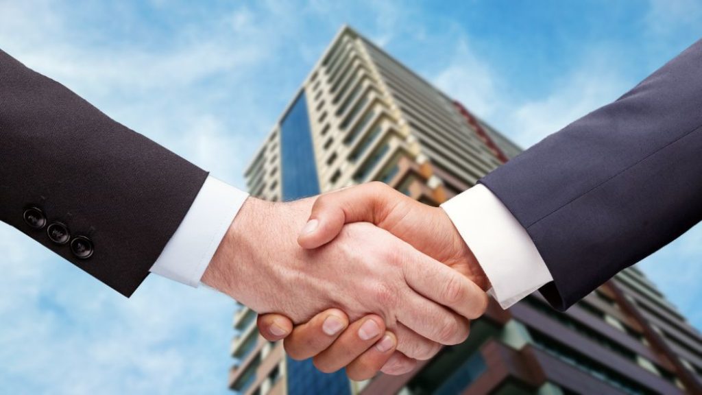Two Professionals Shaking Hands in Front of a Modern Glass Office Building, Symbolizing an Agreement. - Hola Mty
