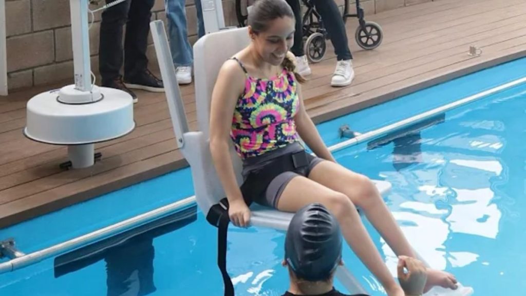 Girl in a Colorful Swimsuit Sits on a Pool Lift Chair at the Edge of a Swimming Pool, with a Caregiver Nearby. - Hola Mty