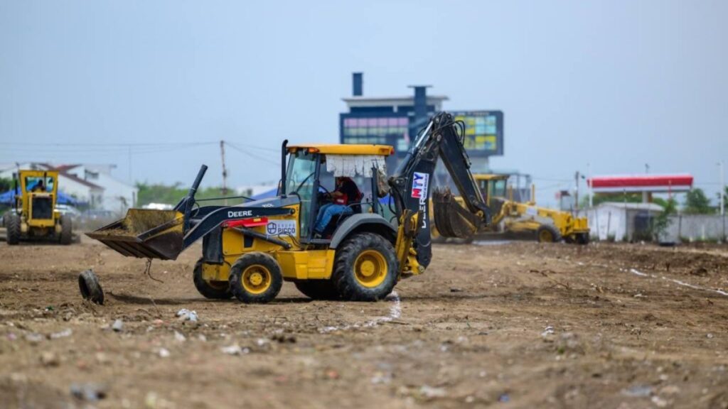 Yellow Backhoe Loader Scooping Dirt on a Cleared Construction Site, with Additional Machinery in the Background and a Billboard Further Away. - Hola Mty