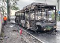 Charred, Burnt-out Bus on a Road with Debris; a Worker in an Orange Shirt Walks Nearby As Emergency Responders Arrive in the Background Behind a Police Vehicle. - Hola Mty