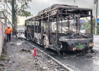 Charred, Burnt-out Bus on a Road with Debris; a Worker in an Orange Shirt Walks Nearby As Emergency Responders Arrive in the Background Behind a Police Vehicle. - Hola Mty