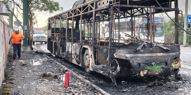 Charred, Burnt-out Bus on a Road with Debris; a Worker in an Orange Shirt Walks Nearby As Emergency Responders Arrive in the Background Behind a Police Vehicle. - Hola Mty