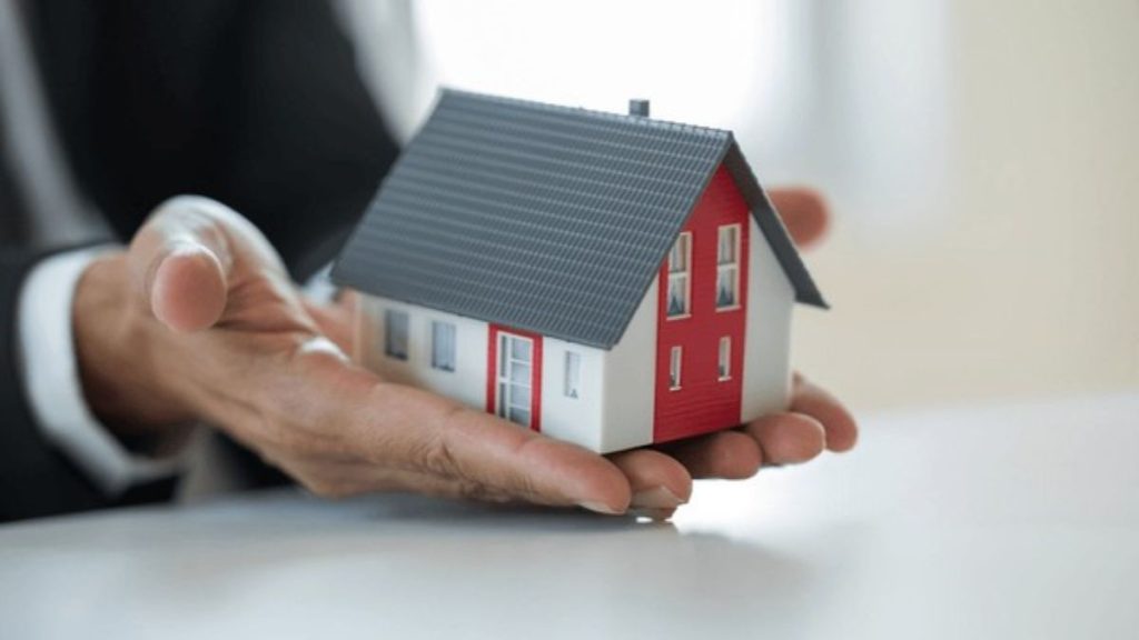 Casa-compra-quantium-desarrollos-nueva-era-inmobiliaria-monterrey - Hola Mty a Person in a Suit Gently Cupping a Small Model House, Symbolizing Real Estate or Home Care. - Hola Mty
