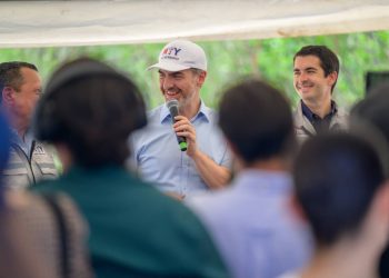 Man in a Light Blue Shirt and White Cap Speaks into a Microphone at an Outdoor Event, Smiling As Colleagues Watch the Crowd. - Hola Mty