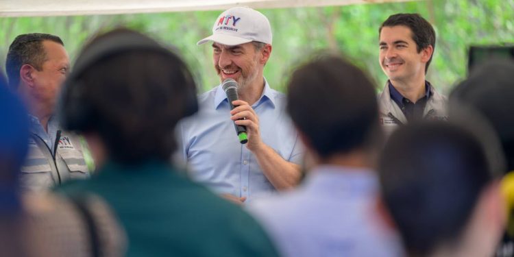 Marcelo-segovia-adrián-de-la-garza - Hola Mty Man in a Light Blue Shirt and White Cap Speaks into a Microphone at an Outdoor Event, Smiling As Colleagues Watch the Crowd. - Hola Mty