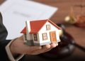 Hands in a Suit Cradle a Miniature White-and-red House Model, with Blurred Legal Desk Items in the Background (papers and Scales). - Hola Mty