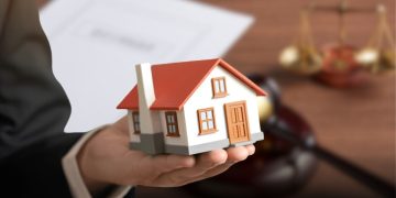 Hands in a Suit Cradle a Miniature White-and-red House Model, with Blurred Legal Desk Items in the Background (papers and Scales). - Hola Mty