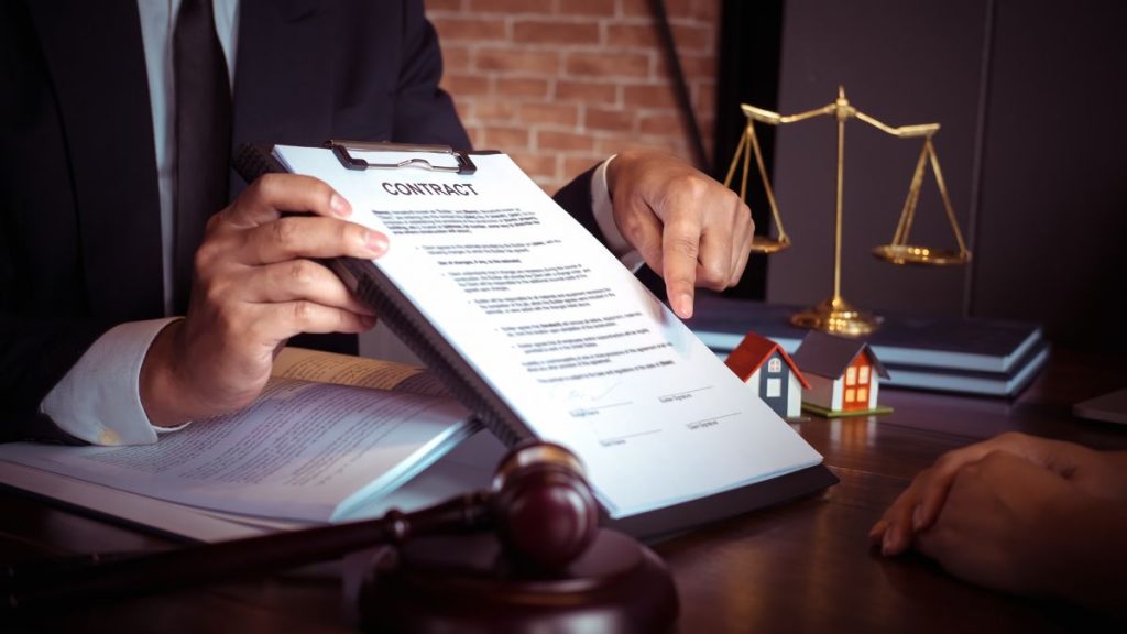 Impuestos-por-comprar-una-vivienda-usada - Hola Mty Lawyer Reviews a Contract on a Desk with a Gavel and Miniature Houses, Scales of Justice in the Background. - Hola Mty