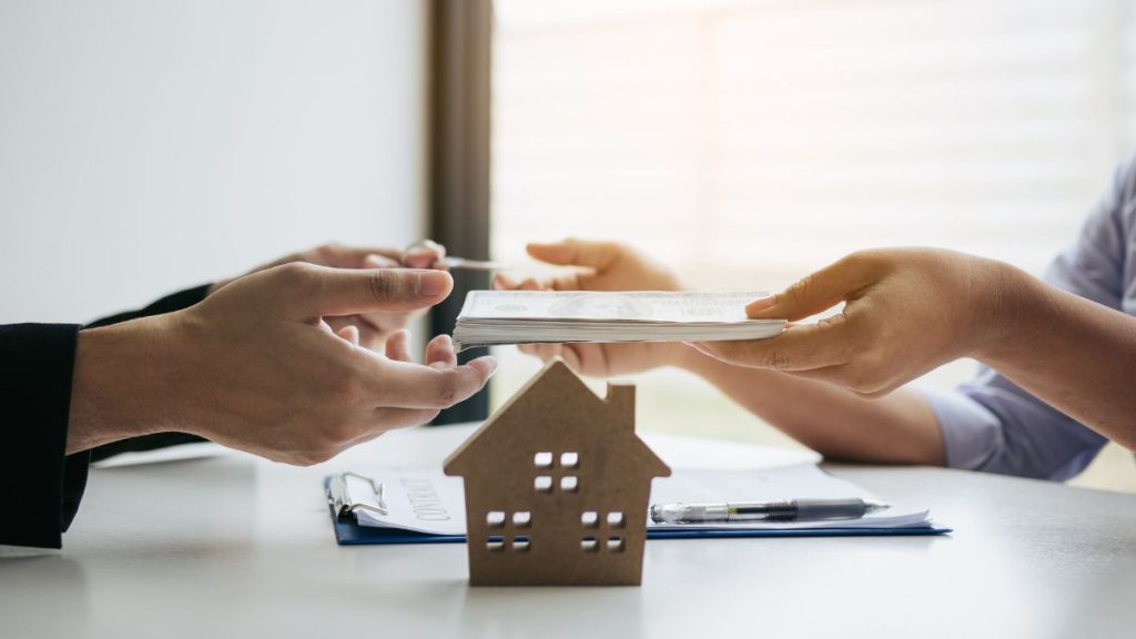 Que-debo-saber-antes-de-comprar-una-vivienda-usada-quantium-desarrollos - Hola Mty Hands Exchanging a Stack of Documents over a Small Cardboard House Model, Symbolizing a Real Estate Deal at a Desk - Hola Mty
